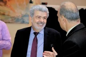Two men are engaged in a conversation, smiling and dressed in formal attire, with a blurred background.