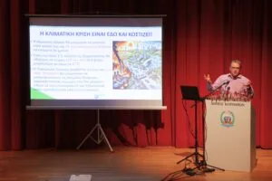 A speaker presents on climate crisis issues in front of a screen displaying information and images, with a podium in a theater setting.