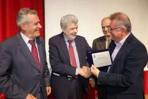 Four men are gathered, smiling and shaking hands. One man is presenting a plaque, while the others look on with approval. Red backdrop.