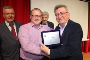 Four men are gathered on stage, with one man presenting a certificate to another. The background features red curtains.