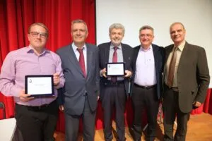 Five men stand together, smiling, holding tablets. They are in front of a red curtain, dressed in formal attire.