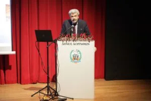A speaker stands at a podium with a logo, addressing an audience in a theater with red curtains. A laptop is nearby.