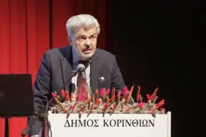 A man speaks at a podium adorned with flowers, in front of a red curtain. The podium has Greek text.