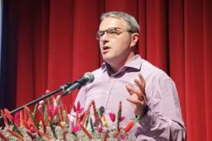 A man in glasses speaks at a podium adorned with colorful flowers, set against a red curtain backdrop.