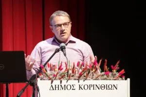 A man in a pink shirt speaks at a podium adorned with flowers, with a red curtain in the background.