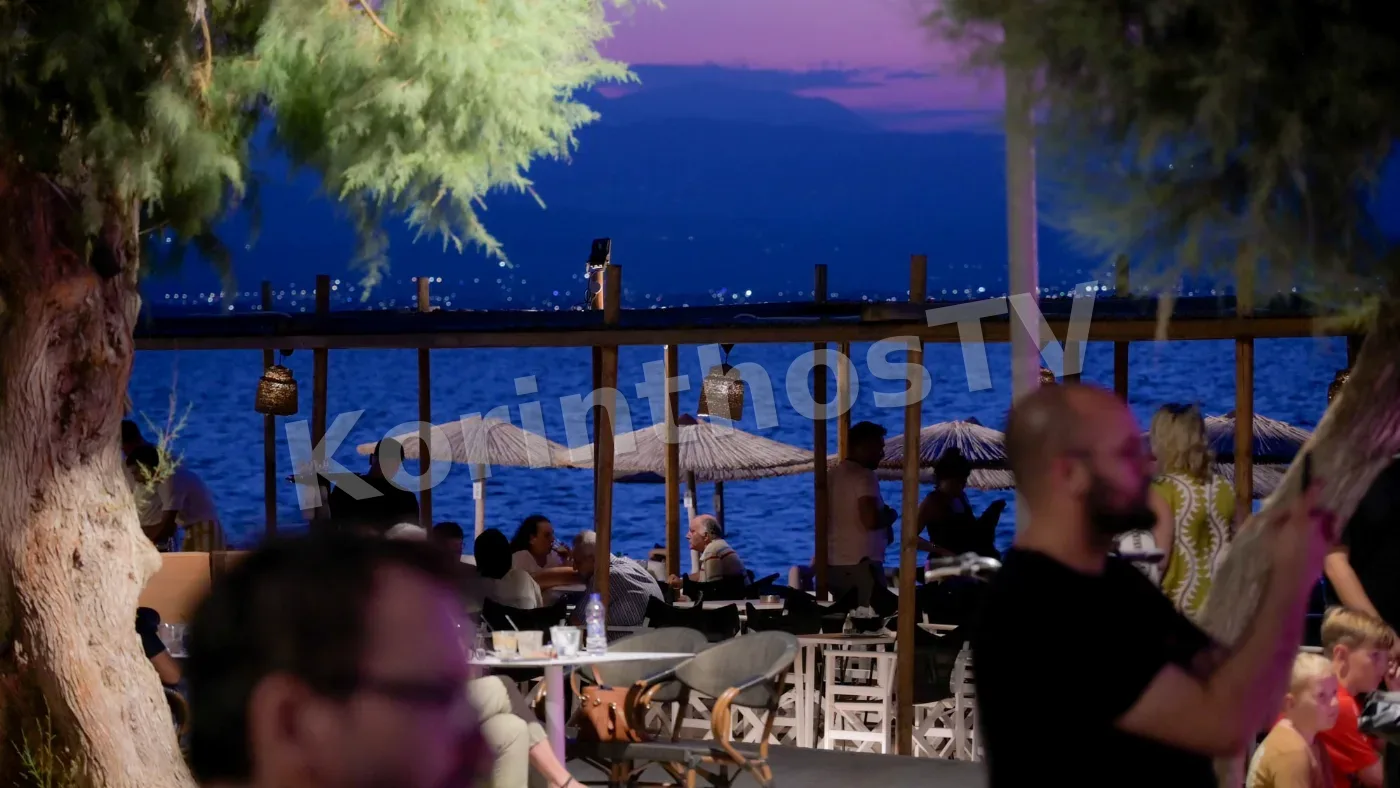 A seaside restaurant scene at dusk, featuring patrons dining under straw umbrellas with a view of the water and distant lights.